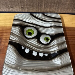 May include: A white glass dish with a mummy face design. The mummy has green eyes, a black mouth, and white teeth. The dish has a textured surface that resembles bandages.