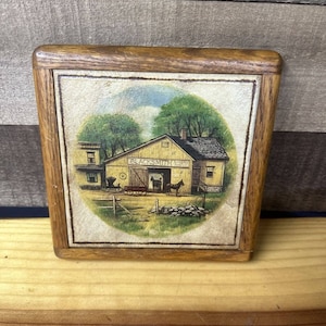 Könnte beinhalten: Ein gerahmter Druck einer Vintage-Illustration, die eine Schmiede mit einem von Pferden gezogenen Wagen davor zeigt. Die Schmiede ist gelb gestrichen und trägt ein Schild mit der Aufschrift "Blacksmith Shop". Der Druck ist in einen Holzrahmen gefasst.