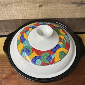 Może przedstawiać: Biały ceramiczny garnek z czarną obwódką i kolorowym geometrycznym wzorem. Garnek ma pokrywkę z gałką na górze.