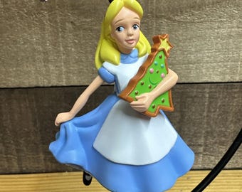 Puede incluir: Un adorno navideño con Alicia de Alicia en el país de las maravillas. La figura lleva un vestido azul y sostiene un árbol de Navidad verde. El adorno tiene una cuerda dorada para colgar.