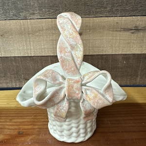 May include: A white ceramic basket with a woven texture, featuring a large bow and handle in shades of pink and yellow. The basket is designed with a decorative bow and handle, perfect for Easter or spring decor.