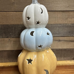 Könnte beinhalten: Gestapelte Kürbisdekoration in Weiß, Hellblau und Gelb. Jeder Kürbis hat Stern- und Halbmond-Ausschnitte. Der obere Kürbis hat einen grauen Stiel. Ein dekorativer Artikel für Herbst oder Halloween.