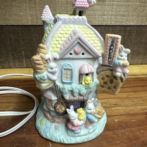 May include: A ceramic Easter-themed house with pastel colors, featuring bunny figurines and a sign that reads "BAKERY." The house has a light-up feature and is decorated with floral and heart accents. A white electrical cord is visible.