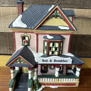 Può includere: Figurina in miniatura di una casa Bed & Breakfast. La casa ha una facciata rosa, un tetto grigio scuro con accenti di neve e un portico con la scritta "Bed & Breakfast". Le rifiniture sono rosse, verdi e gialle. È visibile una piccola ghirlanda natalizia.