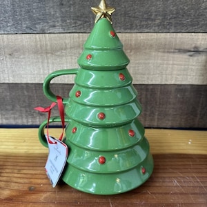 Pode incluir: Uma caneca verde de cerâmica em forma de árvore de Natal, com uma estrela dourada e pontos vermelhos. A caneca tem uma alça e uma tampa a condizer. Uma fita vermelha e uma etiqueta estão presas à alça.