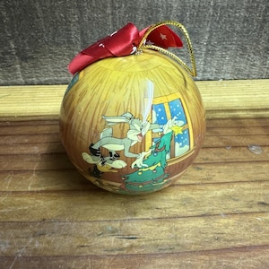 May include: A round Christmas ornament with a Bugs Bunny cartoon scene. The ornament has a red bow and gold string for hanging. The cartoon shows Bugs Bunny decorating a Christmas tree by a window.
