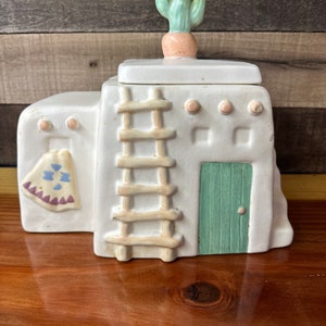 Pode incluir: Cofrinho de cerâmica branca em forma de casa pueblo com uma porta verde, uma escada e um cacto no telhado. A casa tem um cobertor com padrões marrons e brancos pendurado no lado.