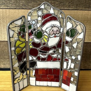 May include: A three-panel stained glass mosaic depicting Santa Claus climbing down a chimney. The mosaic is made of small pieces of glass in various colors, including red, green, white, and gold. The mosaic is framed in silver metal.