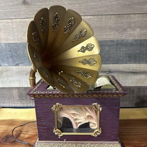 Könnte beinhalten: Eine antike Holz-Spieluhr mit einem verzierten, goldfarbenen Hornlautsprecher. Die Spieluhr hat ein Glasfenster, das einen Papierrollen-Schriftzug im Inneren zeigt. Die Spieluhr steht auf einer Holzoberfläche.