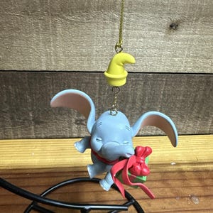 Puede incluir: Adorno azul de Dumbo con orejas grandes, un lazo rojo y un sombrero amarillo. El adorno está suspendido por una cadena dorada. Es un objeto de colección con temática de Disney.