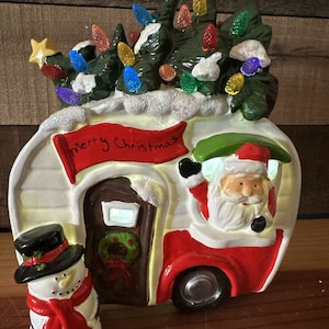 Puede incluir: Una figura de cerámica navideña de una caravana con un muñeco de nieve y Papá Noel. La caravana tiene un techo rojo, lados blancos y una puerta marrón. La caravana tiene un árbol de Navidad en la parte superior con luces de colores. El muñeco de nieve lleva un sombrero de copa negro y una bufanda roja. Papá Noel lleva un gorro rojo y una barba blanca. La caravana tiene una pancarta que dice "Feliz Navidad".