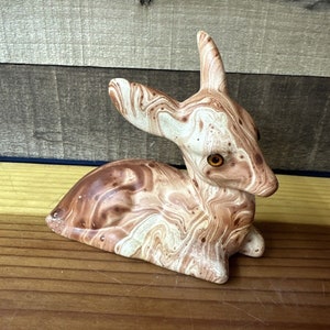 May include: A ceramic fawn figurine with brown and white marbled paint. The fawn is sitting with its head turned to the side and has brown eyes.