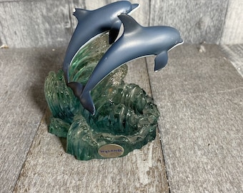 May include: A blue and green resin figurine of two dolphins leaping out of the water. The dolphins are in mid-air, with their tails and fins extended. The base of the figurine is made of clear resin and features a wave-like design. The figurine is signed "Wyland" on the base.