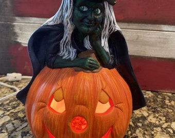 May include: A ceramic witch figurine with green skin and white hair sits on top of a lit-up pumpkin. The witch is wearing a black hat and a black robe. The pumpkin has a carved face with a smiling mouth and two eyes.