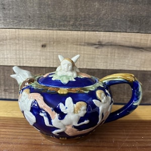 May include: A ceramic teapot with a blue base and lid, featuring white cherubs and floral accents. The handle and spout are cream-colored, with gold and brown details. A cherub figurine sits atop the lid.
