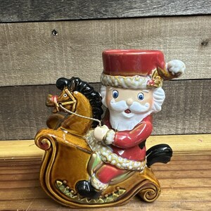 May include: A ceramic Christmas decoration featuring Santa Claus riding a rocking horse. The Santa figure wears a red suit and hat with white trim. The rocking horse is brown with black accents. A festive holiday decoration.