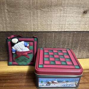 May include: A decorative red and green tin box with a winter scene illustration and a checkerboard pattern on the lid. Beside it is a quilted ornament featuring a doll in a green pocket.