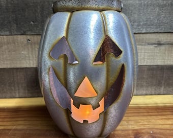 Op de afbeelding: Een zilverkleurige pompoenvormige lantaarn met een uitgesneden jack-o'-lantern gezicht. De lantaarn heeft een roestbruine rand en het gezicht gloeit met een oranje licht. De lantaarn is van metaal en is ontworpen voor Halloween.