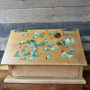 Può includere: Una scatola di legno con un coperchio incernierato dipinto con un motivo di foglie verdi e bacche rosse. La scatola ha uno stile rustico e da fattoria.