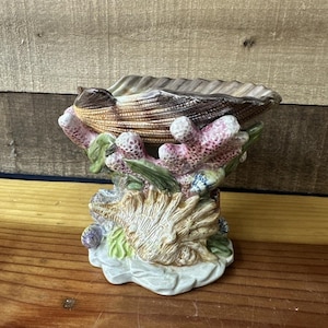 May include: A ceramic seashell-shaped dish with a detailed coral and seashell base. The dish is a light brown color, with pink and white coral accents. The base features a detailed seashell design and is on a white base.