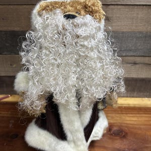 May include: A soft toy dog resembling Santa Claus, with a curly white beard and brown and tan fur. It is wearing a brown and white coat. A festive decorative item.