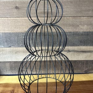 May include: Black metal decorative sculpture composed of three stacked spherical shapes. The sculpture is made of thin metal rods, creating an open, cage-like design. The sculpture is displayed on a wooden surface.