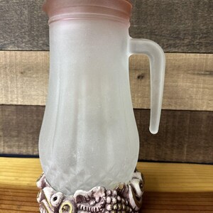 May include: A frosted glass pitcher with a pink rim and a decorative base featuring grapes, leaves, and gold accents.