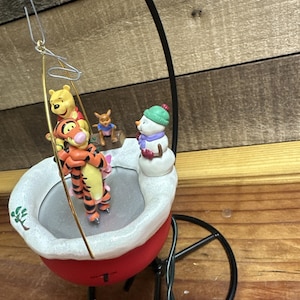 Peut inclure: Ornement décoratif avec les personnages de Winnie l'Ourson. Tigrou, Winnie et Petit Gourou sont sur une balançoire, avec un bonhomme de neige et un petit personnage de Bourriquet à proximité. L'ornement a une base rouge et un bord blanc.
