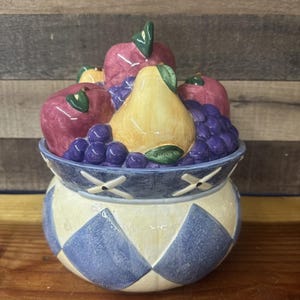 May include: A ceramic fruit bowl with a lid, featuring a colorful assortment of sculpted fruit. The bowl is decorated with blue and white diamond patterns and a blue rim. The fruit includes red apples, purple grapes, and a yellow pear.