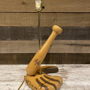 May include: A baseball-themed table lamp featuring a wooden baseball bat and a baseball glove base. The lamp has a light bulb at the top and a brass-colored stem. The baseball bat has a logo on it. The base is shaped like a baseball glove.