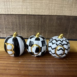 Könnte beinhalten: Drei dekorative Keramik-Kürbisse mit goldenen Stielen, die "BOO" buchstabieren. Jeder Kürbis hat ein anderes schwarz-weißes Muster: Streifen, Schachbrettmuster und Zickzack. Die Buchstaben sind goldfarben.