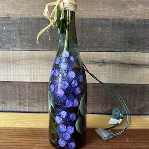 Puede incluir: Una botella de vino de vidrio con un diseño pintado de uvas moradas y hojas verdes. La botella tiene una tapa envuelta en paja y está conectada a un cable de luz con un enchufe.