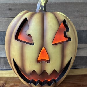 Może przedstawiać: Zabytkowa, podświetlana dekoracja na Halloween w kształcie dyni. Dynia ma klasyczną twarz jack-o'-lantern z trójkątnymi oczami, trójkątnym nosem i poszarpanym uśmiechem. Dynia jest pomalowana w odcieniach żółci i pomarańczy, z zieloną łodygą.