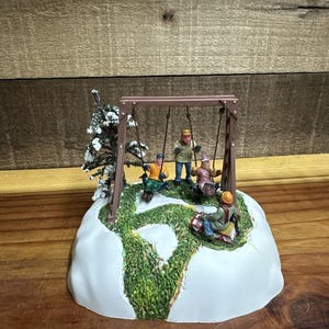 Könnte beinhalten: Eine Miniatur-Winterszene mit einer braunen Schaukel und vier Figuren in Winterkleidung. Die Basis ist mit Schnee und grünem Gras bedeckt. Ein kleiner, schneebedeckter Baum befindet sich links. Winterliches Thema.