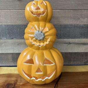 Può includere: Una pila di tre zucche di ceramica a forma di jack-o'-lantern con facce illuminate. Le zucche sono arancioni e hanno un aspetto rustico e vintage.