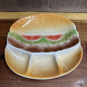 Puede incluir: Una fuente de cerámica con forma de hamburguesa. El pan superior es de color marrón claro con semillas de sésamo, la lechuga es verde y las rodajas de tomate son rojas. La parte inferior tiene tres secciones divididas.