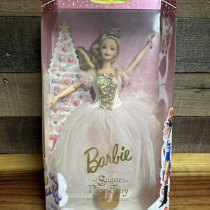 Puede incluir: Muñeca Barbie en una caja de plástico transparente. La muñeca está vestida como el Hada de Azúcar de El Cascanueces, con un corpiño blanco y dorado y un tutú rosa. La caja está etiquetada como "Collector Edition".