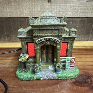 May include: A detailed, illuminated miniature building with the text "CRYPT SWEET CRYPT" above the entrance. The building has a stone-like appearance with red-lit windows. A "Welcome" sign and a "SOLD" sign are also visible.