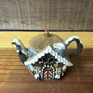 May include: A decorative teapot shaped like a gingerbread house. The teapot features a brown woven lid, a chimney, and a handle. The house has red doors, white trim, and candy accents. Snow-covered roof edges add a festive touch.