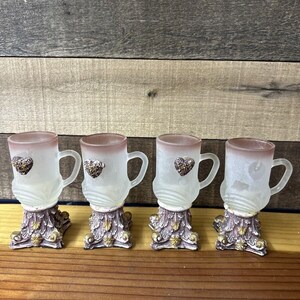 May include: Set of four frosted glass shot glasses with ornate gold and pink bases. Each glass has a small heart design on the front.