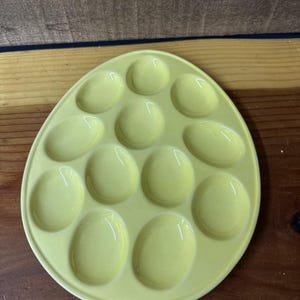 May include: A pale yellow ceramic egg serving tray with 12 egg-shaped indentations. The tray is oval-shaped and has a glossy finish. The tray is designed to hold eggs for serving.