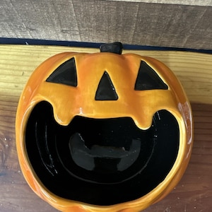 May include: Orange ceramic pumpkin shaped bowl with a black smiling face and a black interior.
