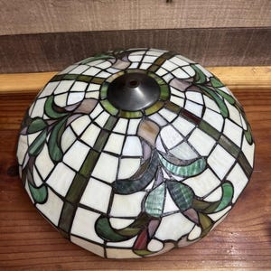 Könnte beinhalten: Ein Buntglas-Lampenschirm mit gewölbter Form. Der Schirm hat ein Muster aus weißen, grünen und braunen Glasteilen, die in einem Gitter- und Blumendesign angeordnet sind. Oben befindet sich eine dunkle Metallkappe.