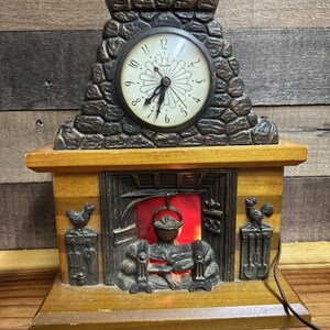 May include: A decorative fireplace clock with a round clock face and a stone-like facade. The clock has a light-up fire effect and rooster and hen accents. The clock is made of wood and metal.