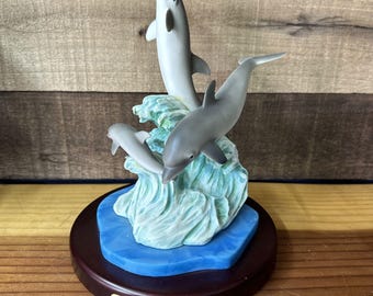 May include: A porcelain figurine of three gray dolphins leaping out of a blue and green wave. The dolphins are in a playful pose, with one dolphin jumping over the other two. The figurine is on a wooden base with a gold plaque that reads "The Gray Rock Collection."