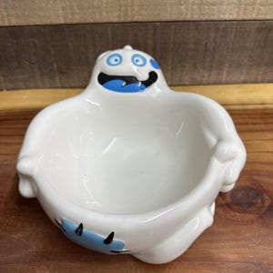 May include: A white ceramic ghost-shaped bowl with a blue and black painted face and accents. The bowl has a wide opening and is designed with rounded edges and a playful, cartoonish style.