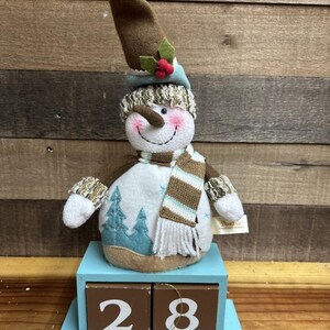 May include: A snowman figurine with a brown hat, scarf, and mittens, standing on a blue wooden block calendar displaying the number 28. The snowman has a smiling face, a carrot nose, and a white body with a winter scene.