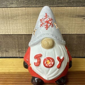 May include: A ceramic gnome figurine with a white hat and red coat. The hat has a snowflake design and the coat has the word "JOY" written on it.