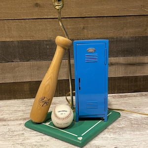 May include: A baseball-themed lamp with a wooden baseball bat base, a baseball, and a small blue locker. The lamp sits on a green base designed to look like a baseball field. The baseball is labeled "CHAMPRO CBH-10 Official League."