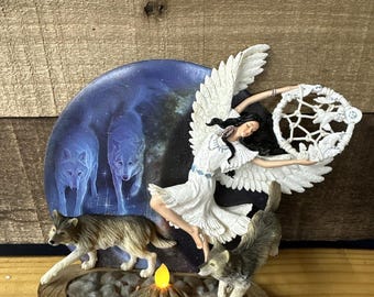 May include: A decorative figurine featuring a woman with angel wings and a dreamcatcher, surrounded by wolves. The background depicts wolves in a starry night scene. The base includes a small, lit candle.